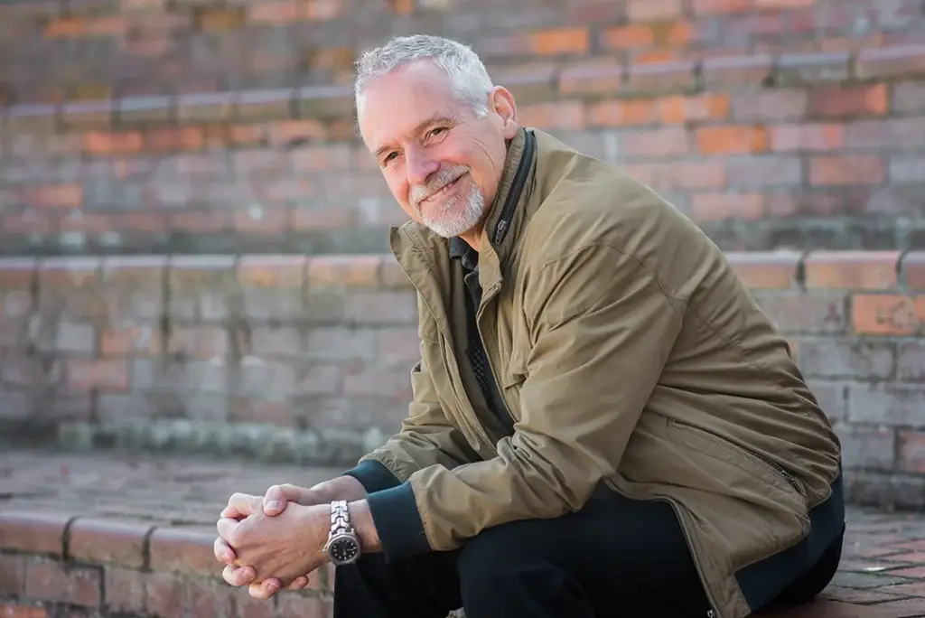 Chris Freer - smiling with grey hair and a beard, wearing a brown jacket and black trousers, sits on outdoor brick steps, smiling warmly at the camera.