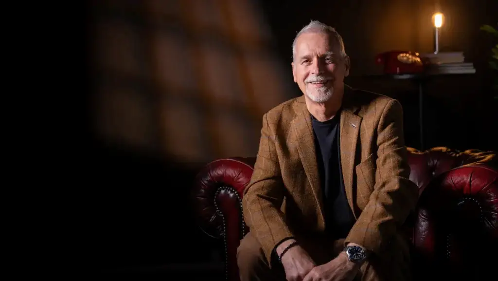 A smiling older man with short grey hair and a beard, wearing a brown blazer and sitting on a red leather sofa. A lit lamp, red telephone, and books are in the softly lit background.