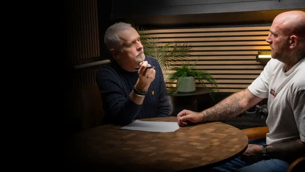Two men sit at a round wooden table indoors, engaged in a serious conversation. One man gestures with a pen, holding a paper, while the other listens. A potted plant and striped wooden wall are in the background.