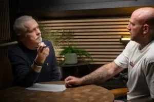 Two men sit across a table, one older with grey hair holding a pen and paper, and the other bald with tattoos, engaged in a serious conversation. A potted plant and a light are visible in the background.