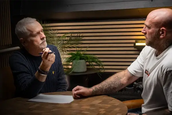 Two men sit across a table, one older with grey hair holding a pen and paper, and the other bald with tattoos, engaged in a serious conversation. A potted plant and a light are visible in the background.