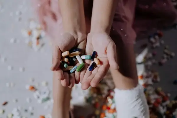 A person holds various colourful pills and capsules in their hands, with more scattered on the floor below. The person appears to have an injured foot wrapped in a bandage.