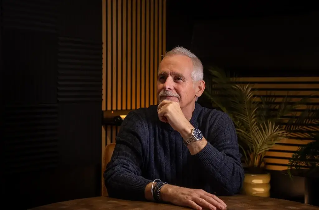 An older man with grey hair and a beard sits at a table, resting his chin on his hand and smiling thoughtfully. He is wearing a dark jumper and a watch. The background features plants and vertical wooden slats.