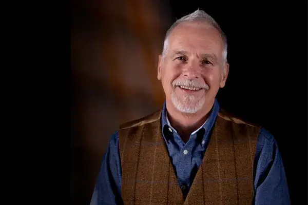 An older man with short white hair and a goatee smiles at the camera. He is wearing a blue button-up shirt and a brown checked waistcoat, standing against a dark, softly blurred background.