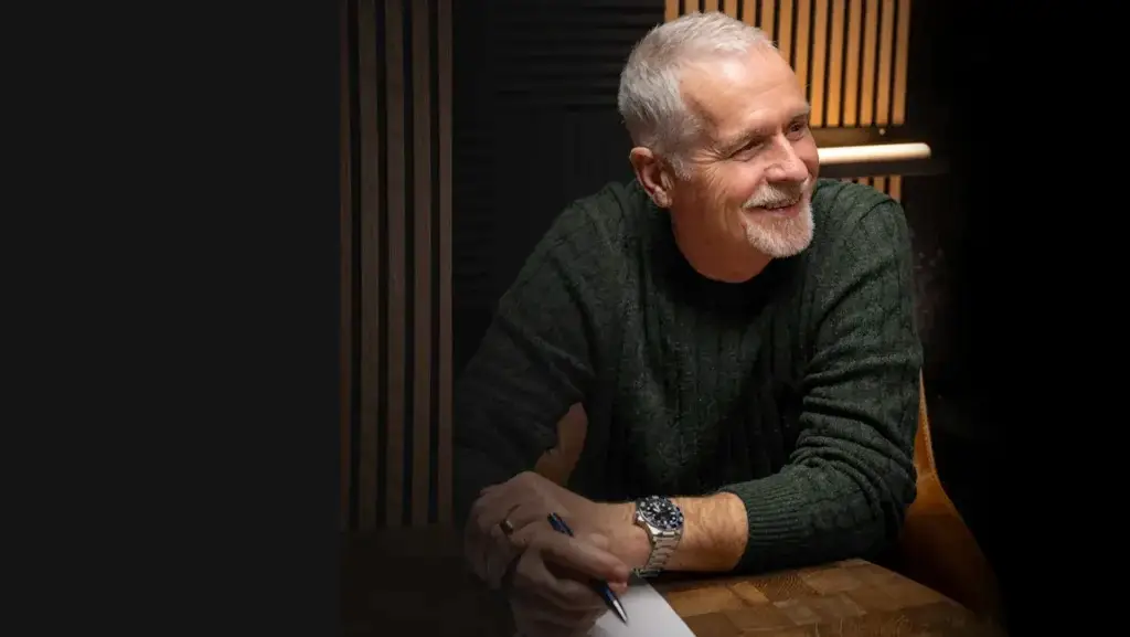 An older man with short grey hair and a beard, wearing a green jumper and watch, smiles whilst sitting at a wooden table holding a pen and paper. Warm lighting and vertical wooden panels are in the background.
