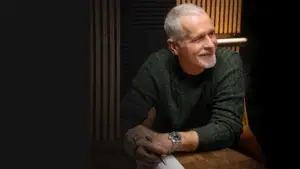 An older man with short grey hair and a beard, wearing a green jumper and watch, smiles whilst sitting at a wooden table holding a pen and paper. Warm lighting and vertical wooden panels are in the background.