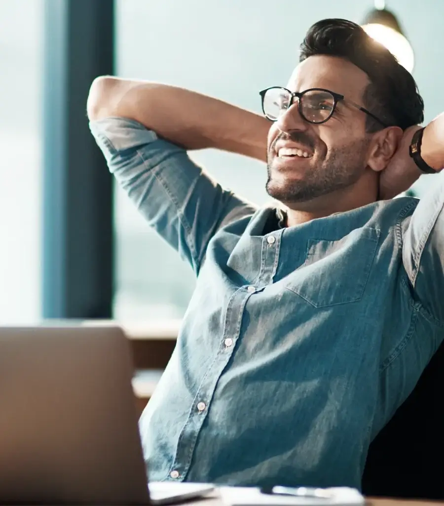 A man wearing glasses and a denim shirt leans back in his chair, smiling with his hands behind his head, in front of a laptop in a bright, modern office space.