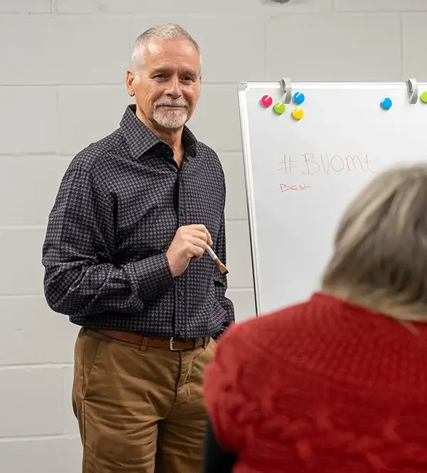 An older man with grey hair and a beard stands by a whiteboard, holding a marker and smiling at a seated person in a red jumper. The whiteboard has “#BV0mt” written on it.