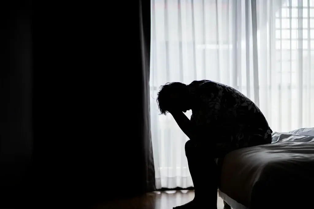A person sits on the edge of a bed in a dark room, hunched over with their head in their hands, silhouetted against a window with net curtains, conveying a sense of sadness or distress.