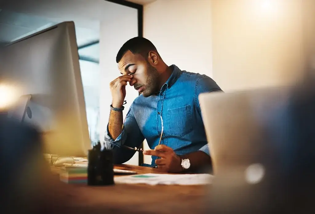 A man sitting at a desk in front of a computer holds his glasses in one hand and pinches the bridge of his nose with his other hand, appearing stressed or tired whilst working.