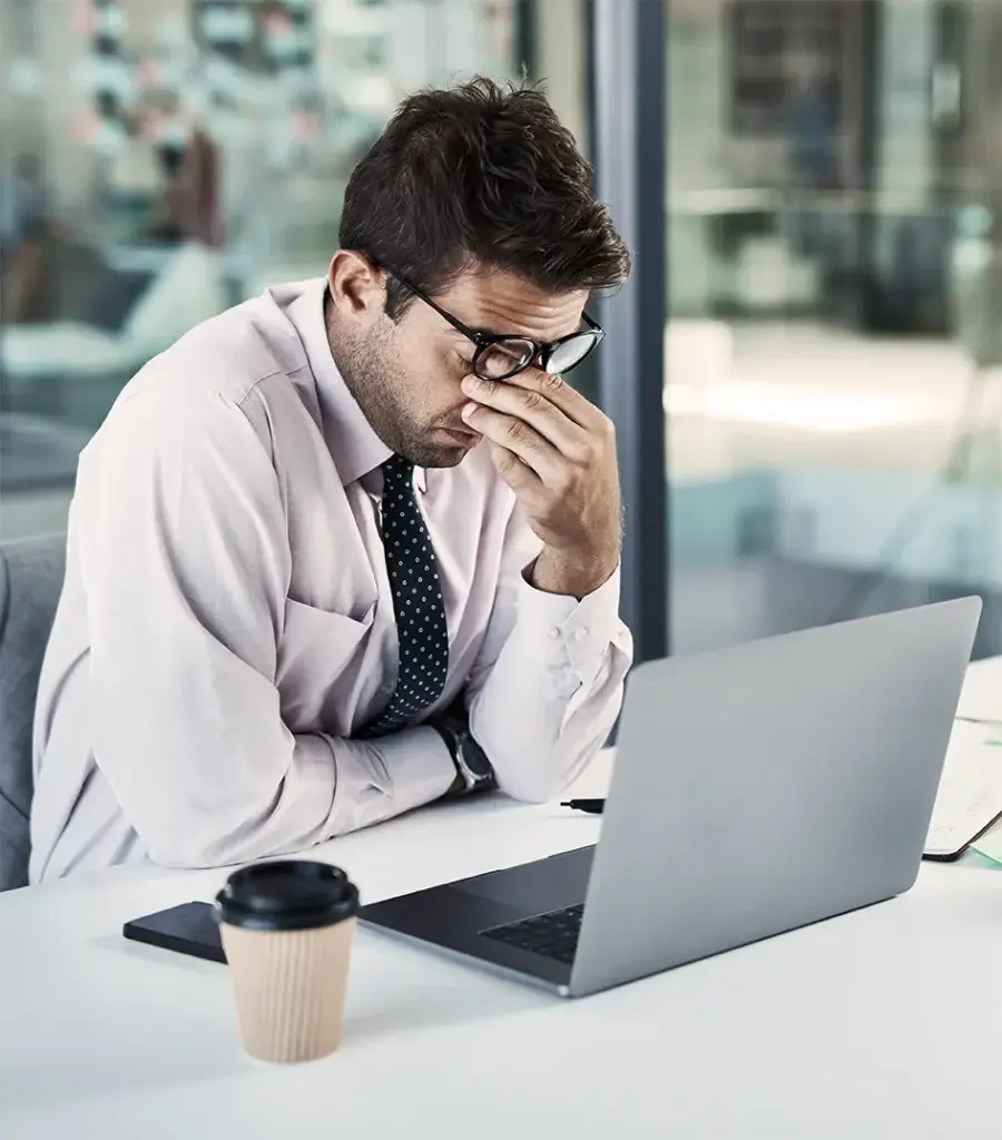 A man in a shirt and tie sits at a desk with a laptop, holding his glasses and pinching the bridge of his nose, appearing stressed or tired. A coffee cup and documents are on the desk.
