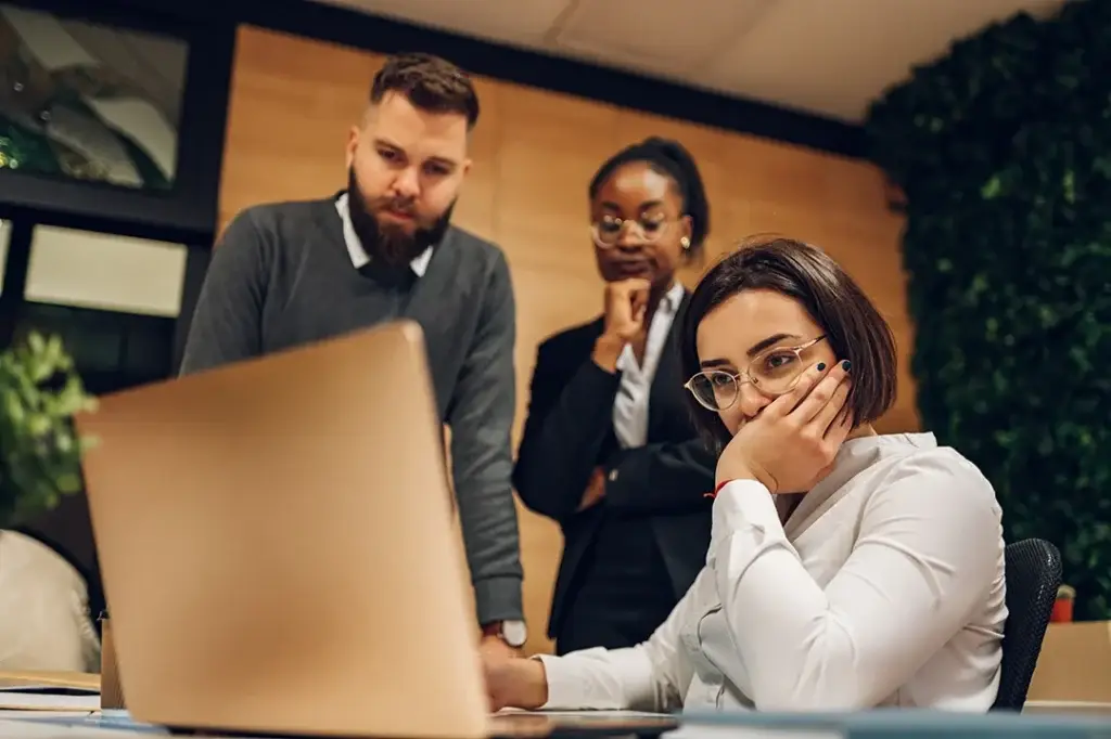 Three concerned colleagues stand around a seated woman looking at a laptop screen. The woman has her hand over her mouth, whilst the others appear thoughtful and serious in an office setting.
