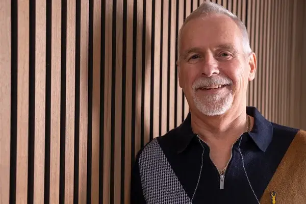 A smiling older man with grey hair and a white beard stands against a brown wooden slat wall, wearing a dark zip-up jumper with a geometric pattern and gold accents.