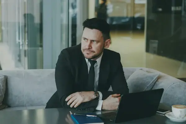 A man in a black suit sits at a table with a laptop, notebook, and coffee cup, looking thoughtfully to the side in a modern office setting.
