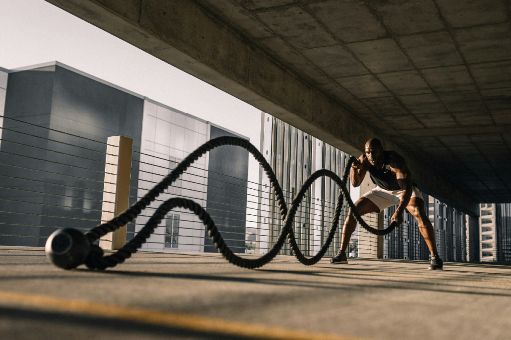 A man in sportswear trains with battle ropes in an outdoor urban setting under a concrete roof, creating waves with the ropes while maintaining a powerful, focused stance.