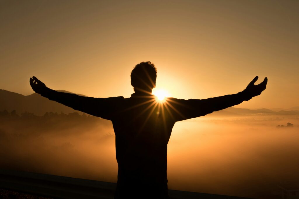 A person stands with arms outstretched, facing the sunrise over a misty landscape. The sun shines brightly, creating a dramatic silhouette and warm golden light across the scene.