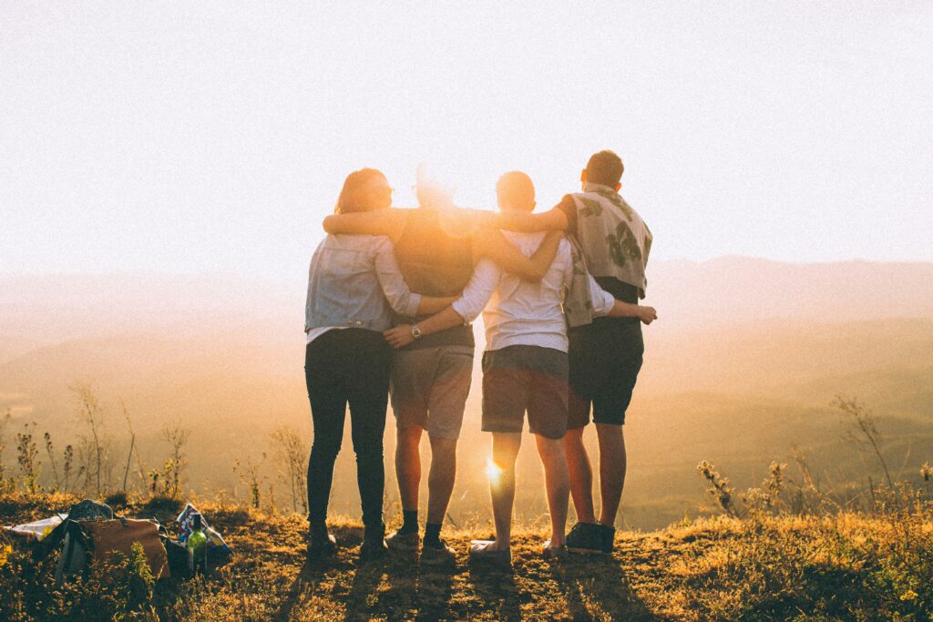 Four people stand together outdoors at sunset, arms around each other, looking at a scenic view of hills and sky. The warm sunlight creates a bright, golden glow around them.