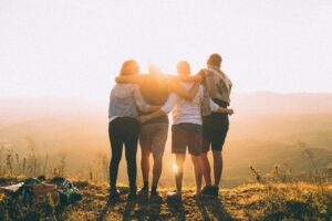 Four people stand together outdoors at sunset, arms around each other, looking at a scenic view of hills and sky. The warm sunlight creates a bright, golden glow around them.