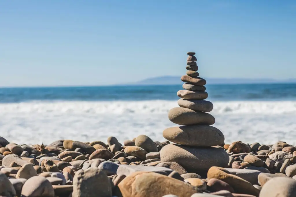 A stack of smooth, rounded stones balanced on a rocky beach with waves crashing in the background and a distant mountain visible under a clear blue sky.