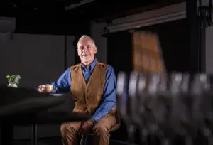 An older man with grey hair and a beard, wearing a brown waistcoat and blue shirt, sits at a table holding a cup of coffee. He smiles at the camera in a dimly lit room with blurred glasses in the foreground.