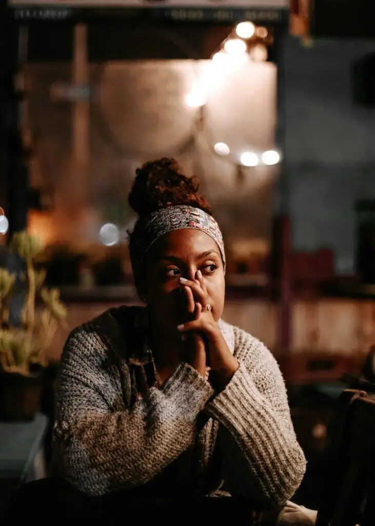 A woman wearing a hairband and a cosy jumper sits indoors with her hand resting on her mouth, looking thoughtfully to the side. Warm, soft lighting creates a calm and contemplative atmosphere around her.