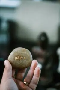 A hand holds a smooth stone with the word Believe engraved in gold letters. The background is blurred, suggesting an indoor setting.